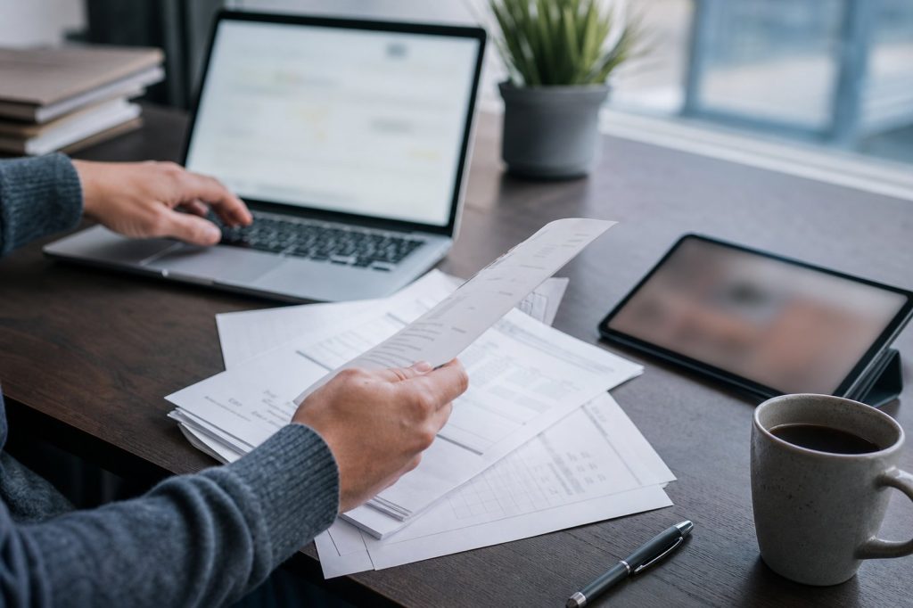 Small-business owner manually copying customer details from paper invoices into a laptop next to a separate tablet